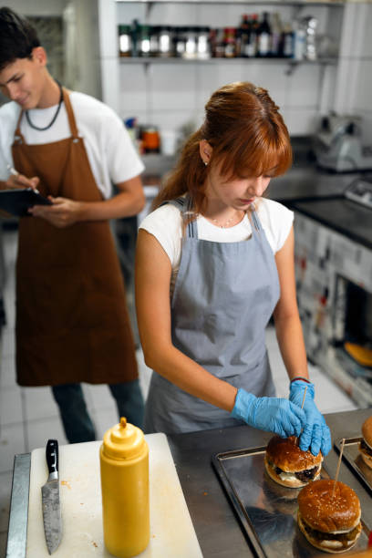 teenage girl making burgers int he fast food restaurant kitchen, while her coworker taking orders - junk food stock pictures, royalty-free photos & images
