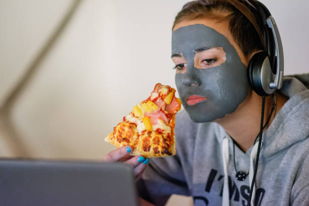 teenage girl wearing headphones and watching her laptop while holding a slice of pizza. - junk food stock pictures, royalty-free photos & images