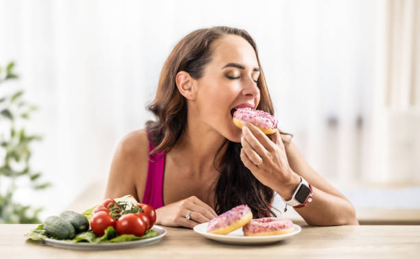 the young woman couldn't resist the sweet temptation, she excitedly bit into a sweet donut and gave preference to healthy vegetables. - junk food stock pictures, royalty-free photos & images