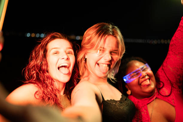 three young women taking selfie sticking out tongues at night - concert stock pictures, royalty-free photos & images
