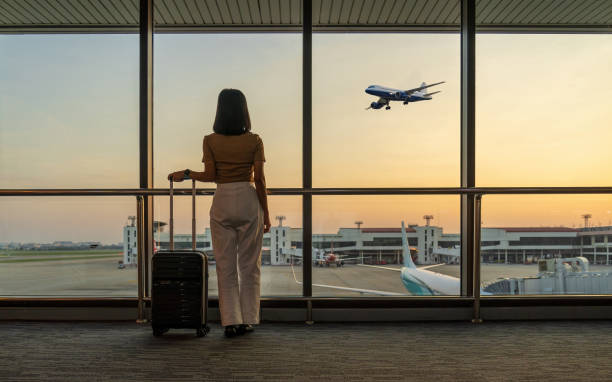 travel tourist standing with luggage watching sunset at airport window. woman looking at lounge looking at airplanes while waiting at boarding gate before departure. travel lifestyle. transport and travel concept - travel sto