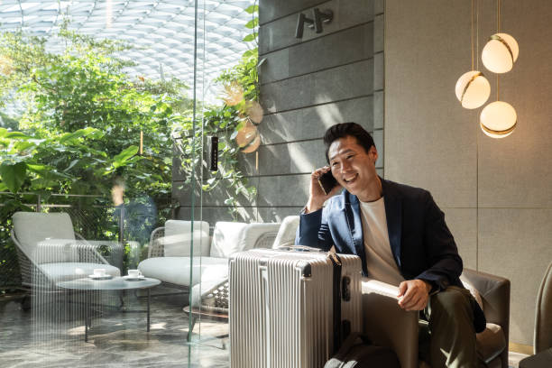 travelling asian businessman smiles at an airport lounge after receiving great news over the phone - travel stock pictures, royalty-free photos & images