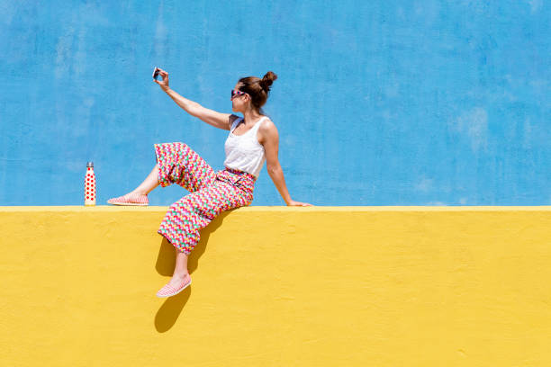 trendy woman sitting on yellow wall while taking a selfie - fashion stock pictures, royalty-free photos & images
