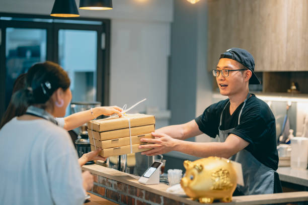 two asian customers come to pick up their take-out pizzas, and the pizza shop owner personally hands over the ordered meals to them. - junk food stock pictures, royalty-free photos & images