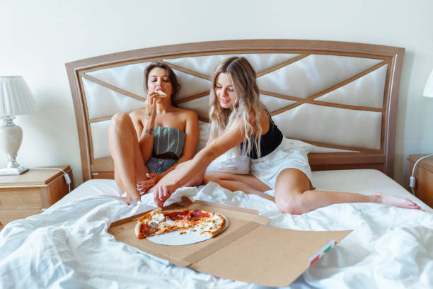 two barefoot women relaxing in a bed in the morning, eating pizza, going for the biggest slice - junk food stock pictures, royalty-free photos & images