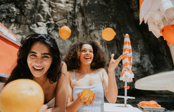 two beautiful woman juggle oranges on a sunny beach - travel stock pictures, royalty-free photos & images