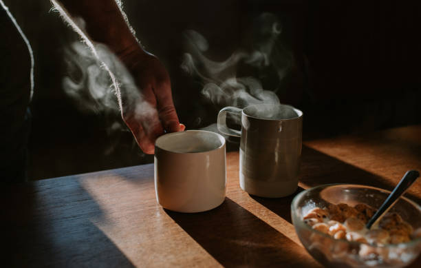 two cups of coffee on a wooden table with a bowl of breakfast cereal. someone lifts one of the mugs out of frame. - coffee stock pictures, royalty-free photos & images