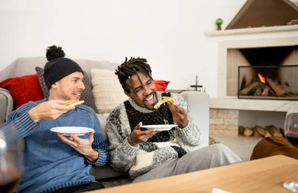 two diverse male friends laughing and eating pizza by a fire in winter - junk food stock pictures, royalty-free photos & images