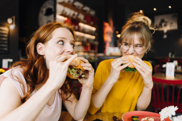 two female friends eating burger in a restaurant - food stock pictures, royalty-free photos & images