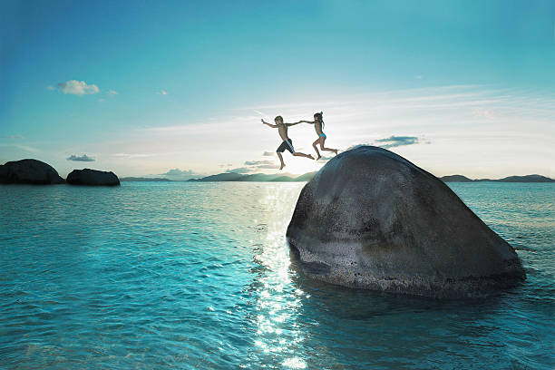 two kids holding hands jumping off rock into sea - travel stock pictures, royalty-free photos & images