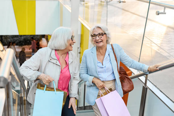 two senior women are walking up the stairs in a shopping mall while having fun shopping - fashion stock pictures, royalty-free photos & images