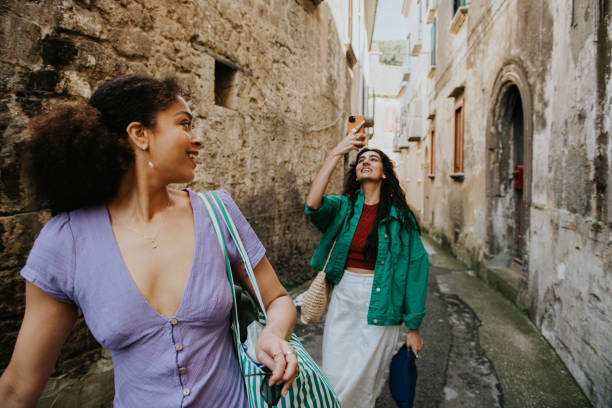 two stylish woman walk down an alleyway. one takes a photo using her mobile phone. - travel stock pictures, royalty-free photos & images