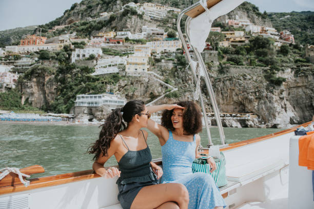 two woman enjoy a journey on a boat, with positano, italy, in the background. one woman wears sunglasses, the other shields her eyes from the bright light with her hands. - travel stock-fotos und bilder