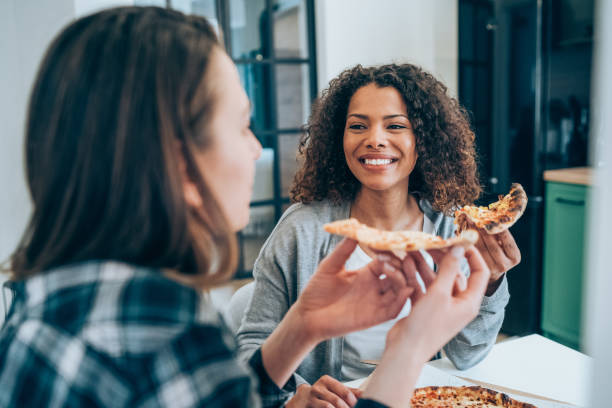 two young women eating pizza together at home. - junk food stock pictures, royalty-free photos & images