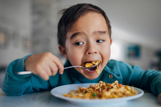 una vista frontal de un niño hambriento de raza mixta comiendo arroz frito para el almuerzo con una cuchara. el fondo está borroso. - food fotografías e imágenes de stock