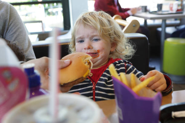 upset toddler being given burger in fast food restaurant - junk food stock pictures, royalty-free photos & images