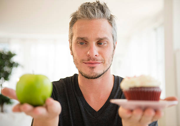 usa, new jersey, jersey city, man weighing green apple against cup cake - junk food stock pictures, royalty-free photos & images