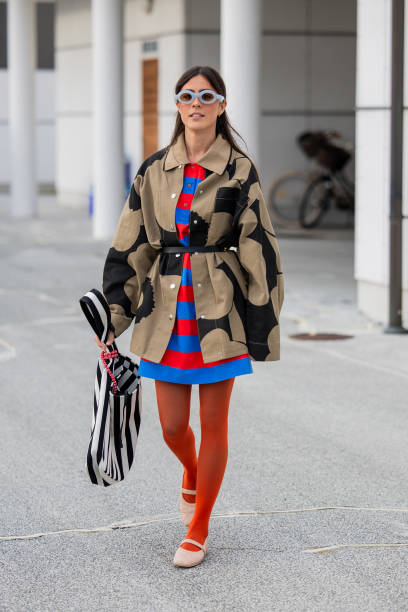 Vicky Montanari wears brown belted jacket, striped blue red dress, red tights, black white striped bag outside Marimekko during the Copenhagen...