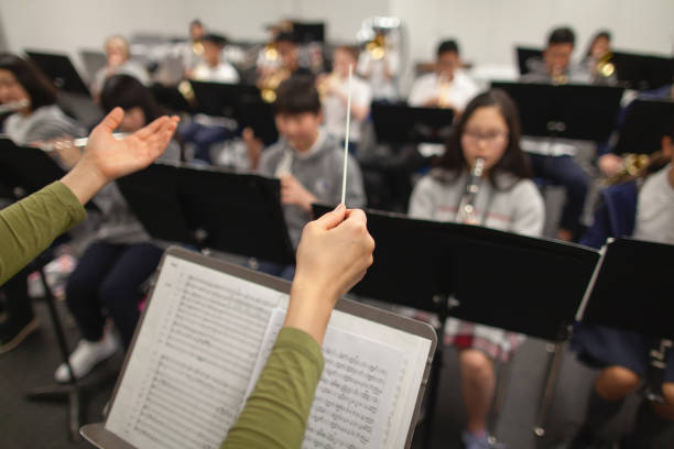 view of band instructor teacher conductor hands leading music performance orchestra of junior high school teenager students practicing in school classroom together in a group of people - concert stock pictures, royalty-free p