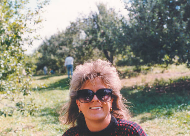 vintage 1980s hairstyle, stylish 1990s hair, happy woman wearing sunglasses outdoors apple picking - hairstyle stock-fotos und bilder