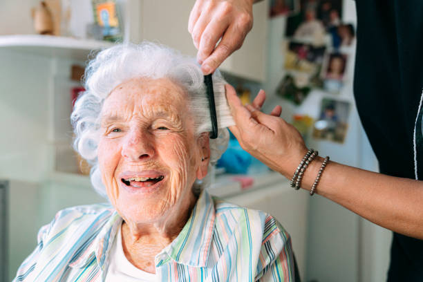 weitwinkelaufnahme einer fröhlichen, niedlichen älteren kaukasischen frau zu hause, die sich von einem professionellen stylisten für häusliche pflegekräfte die haare kämmen lässt - hairstyle stock-fotos und bilder