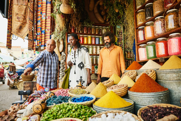 wide shot couple talking to spice shop owner in the souks of marrakech - travel stock pictures, royalty-free photos & images
