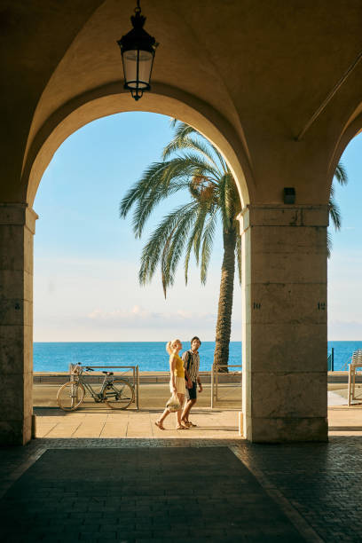 wide shot of a couple on holiday walking under an archway on the promenade des anglais in nice, france - travel stock pictures, royalty-free photos & images