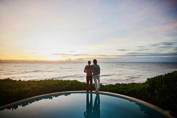wide shot of embracing gay couple watching sunrise while standing at edge of pool at luxury tropical villa overlooking ocean - travel stock pictures, royalty-free photos & images