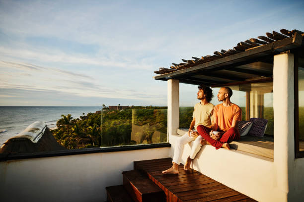 wide shot of gay couple sitting on rooftop deck of luxury tropical beachfront villa while drinking coffee and watching sunrise - travel stock pictures, royalty-free photos & images