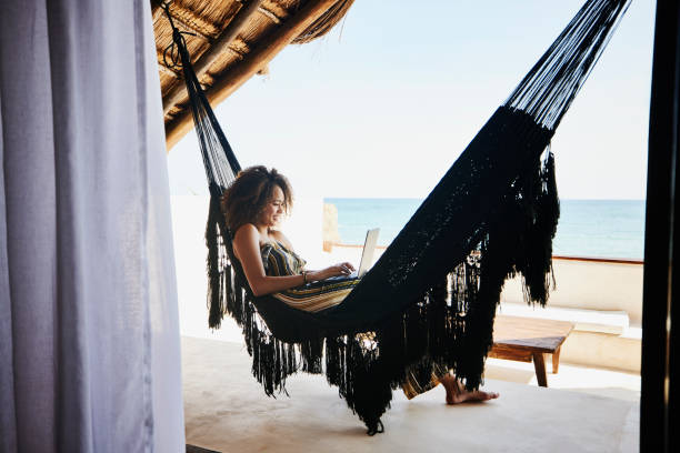wide shot of woman working on laptop while relaxing in hammock on deck of luxury tropical villa overlooking ocean - travel stock pictures, royalty-free photos & images