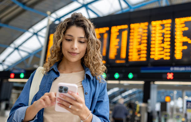 woman at the train station using her phone while checking the departure board - travel stock pictures, royalty-free photos & images