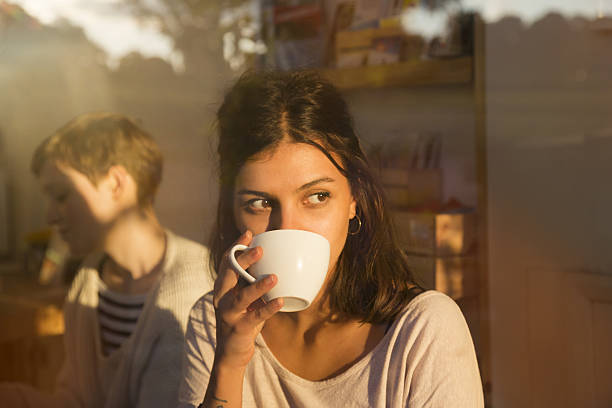 woman drinking coffee in urban cafe while looking outside, sunny reflections in window. - coffee stock pictures, royalty-free photos & images