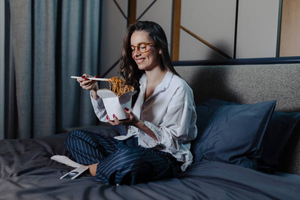 woman eating pasta and watching movie at bed at night - junk food stock pictures, royalty-free photos & images