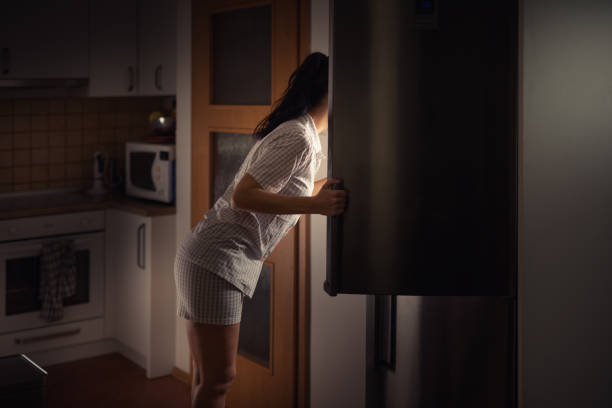 woman leaning into fridge at night searching for food with bad diet habits. - junk food stock pictures, royalty-free photos & images