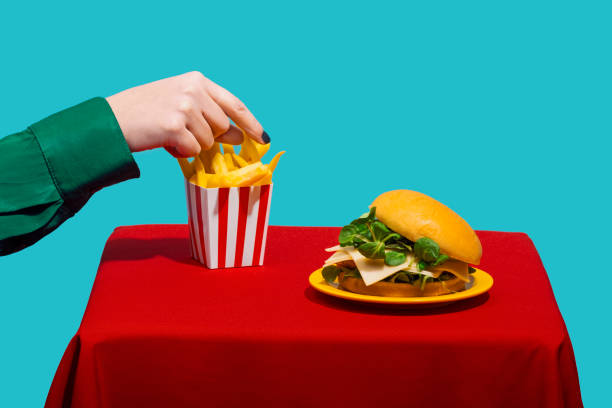 woman reaching for french fries near cheeseburger on table against blue background - junk food stock pictures, royalty-free photos & images