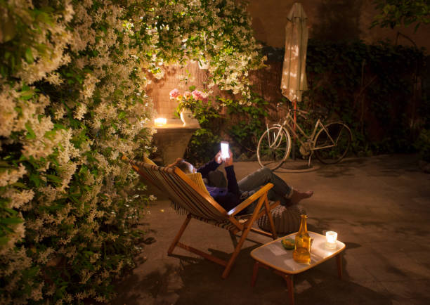 woman relaxing on deck chair in backyard at night, holding mobile phone - garden decoration photos et images de collection