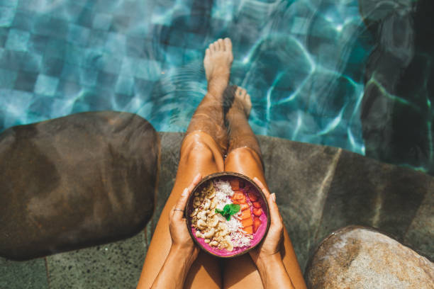 woman's hand holding fresh fruit smoothie bowl on background of turquoise water in swimming pool - food stock pictures, royalty-free photos & images