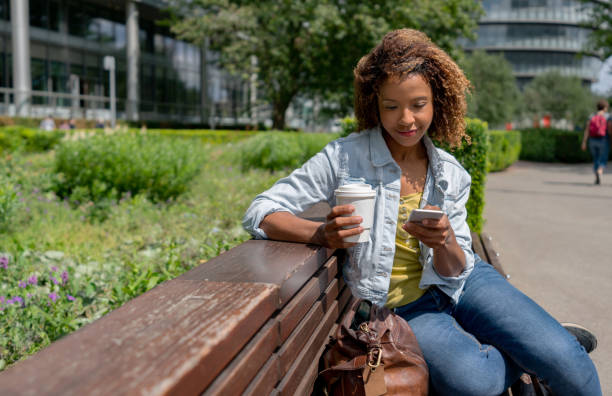 woman sitting on a bench outdoors social networking on her phone - junk food stock pictures, royalty-free photos & images