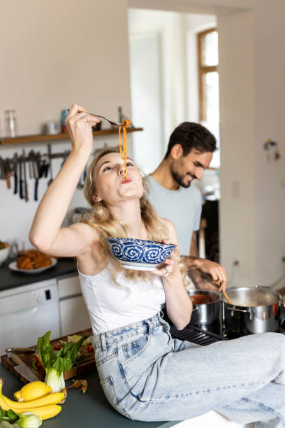 woman slurping noodles with boyfriend preparing food in kitchen at home - food stock pictures, royalty-free photos & images