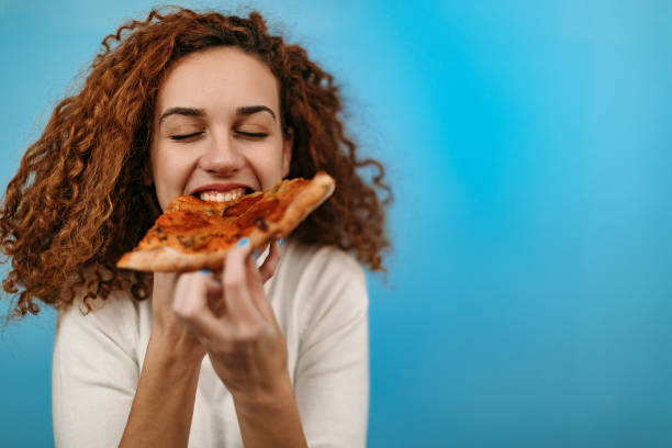 woman standing in front of blue background and eating pizza - junk food stock pictures, royalty-free photos & images