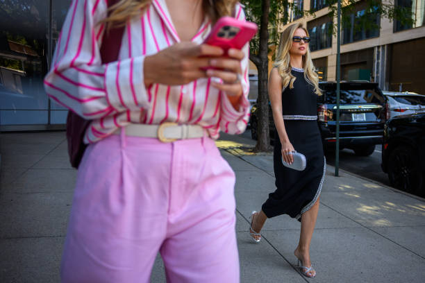 Woman wearing a black dress with silver trim with a silver purse and sandel high heels walks near a woman on her phone outside Pat Bo during the New...