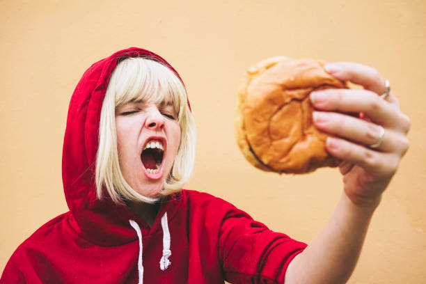 woman with eyes closed holding hamburger in front of peach wall - junk food stock-fotos und bilder
