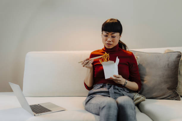 woman working on laptop and eating pasta while sitting on sofa - junk food stock pictures, royalty-free photos & images