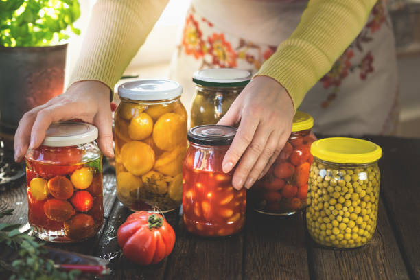 women with traditional apron holding jars with various pickled preserved vegetables on rustic, wooden kitchen table. - food stock pictures, royalty-free photos & images