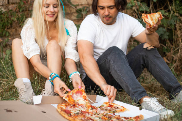 wonderful couple sitting on ground outdoors and taking pieces of fresh pizza from box. - junk food stock pictures, royalty-free photos & images