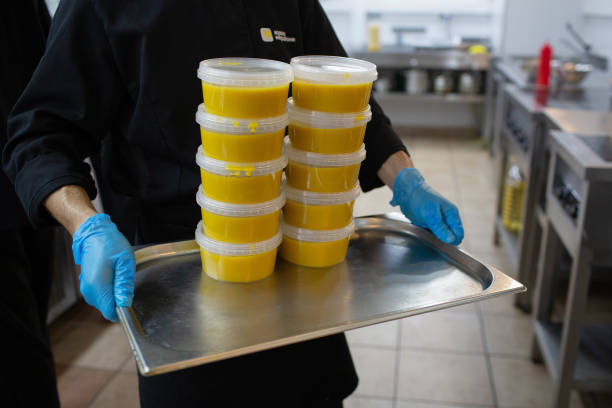 Worker wearing latex gloves carries stacks of soup portions in plastic containers inside a Localkitchen.ru food delivery kitchen in Moscow, Russia,...