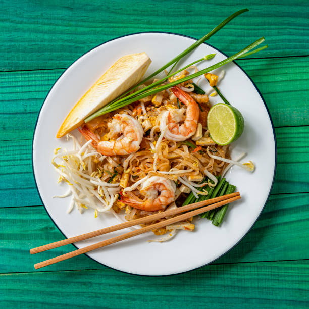 world famous freshly stir-fried cooked, thai recipe of prawn pad thai noodles on a round traditional enameled metal dish with chopsticks laid on the side of the dish, set on an abstract weathered turquoise colored wood panel 