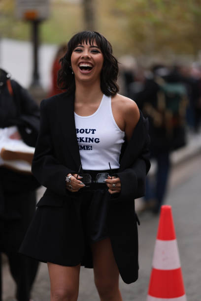 Xochitl Gomez is seen wearing an oversized black blazer with peak lapels and shoulder pads, a white tank top with a dark blue printed quote saying...