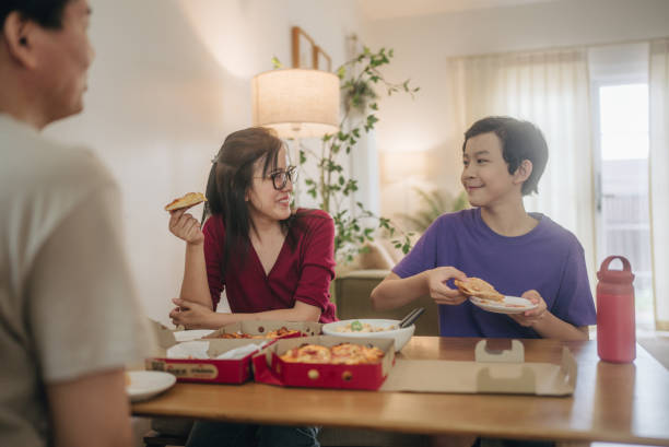 young asian boy sharing his piece of pizza to his mom while they enjoy lunch together on weekend at home. - junk food stock pictures, royalty-free photos & images