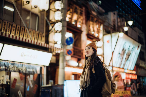 young asian female traveller with backpack visiting japan, exploring and strolling in downtown city street at night in osaka. a popular nightlife and entertainment area in the city. travel, vacation and holiday concept - trav